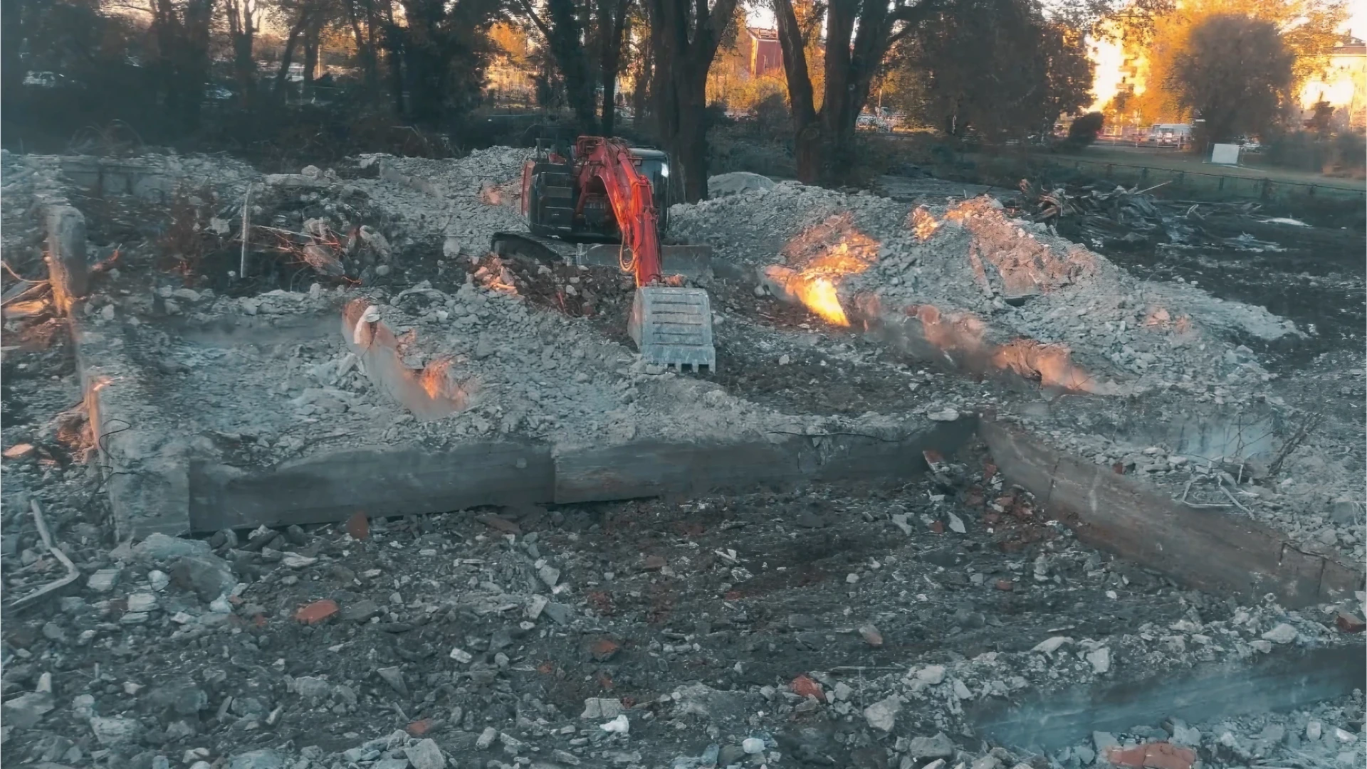 Escavatore rosso su cantiere di demolizione, macerie e fondazioni esposte.