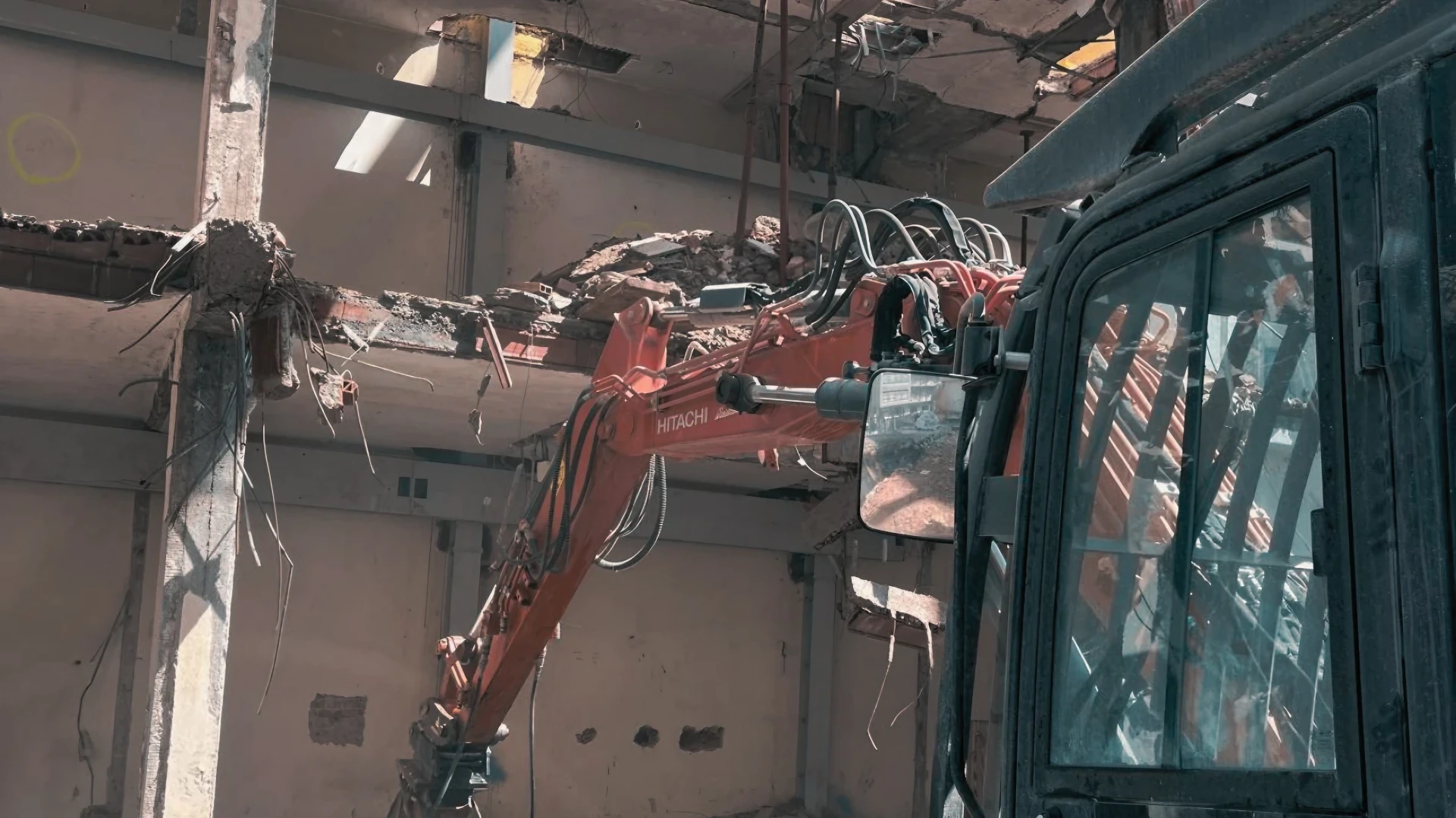 Escavatore Hitachi in azione per demolizione controllata di struttura interna, con detriti e ferri esposti.